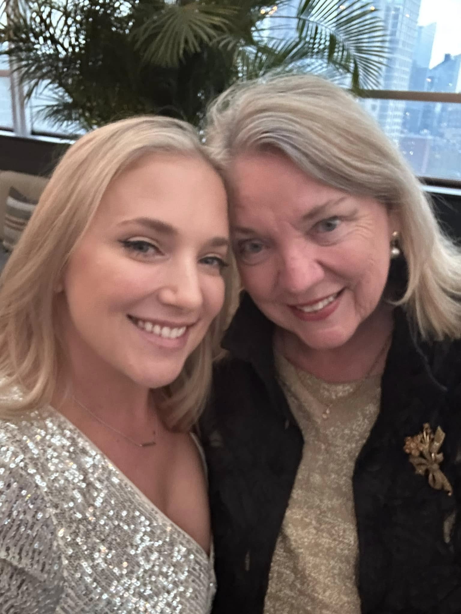 Ellen and her mother at a rooftop restaurant with a city skyline behind them. Ellen wears a silver sequined top; her mother has silver-white hair and pearl earrings. Both dressed up, heads together.