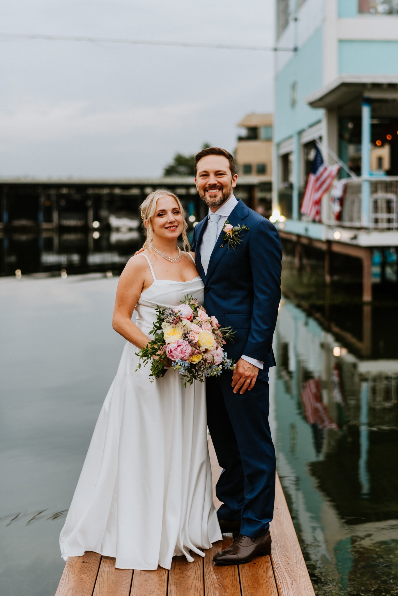 Ellen in a white A-line gown holding a bouquet of pink, yellow, and blue flowers, standing on a wooden dock at dusk with her husband in a navy suit. Both smiling.