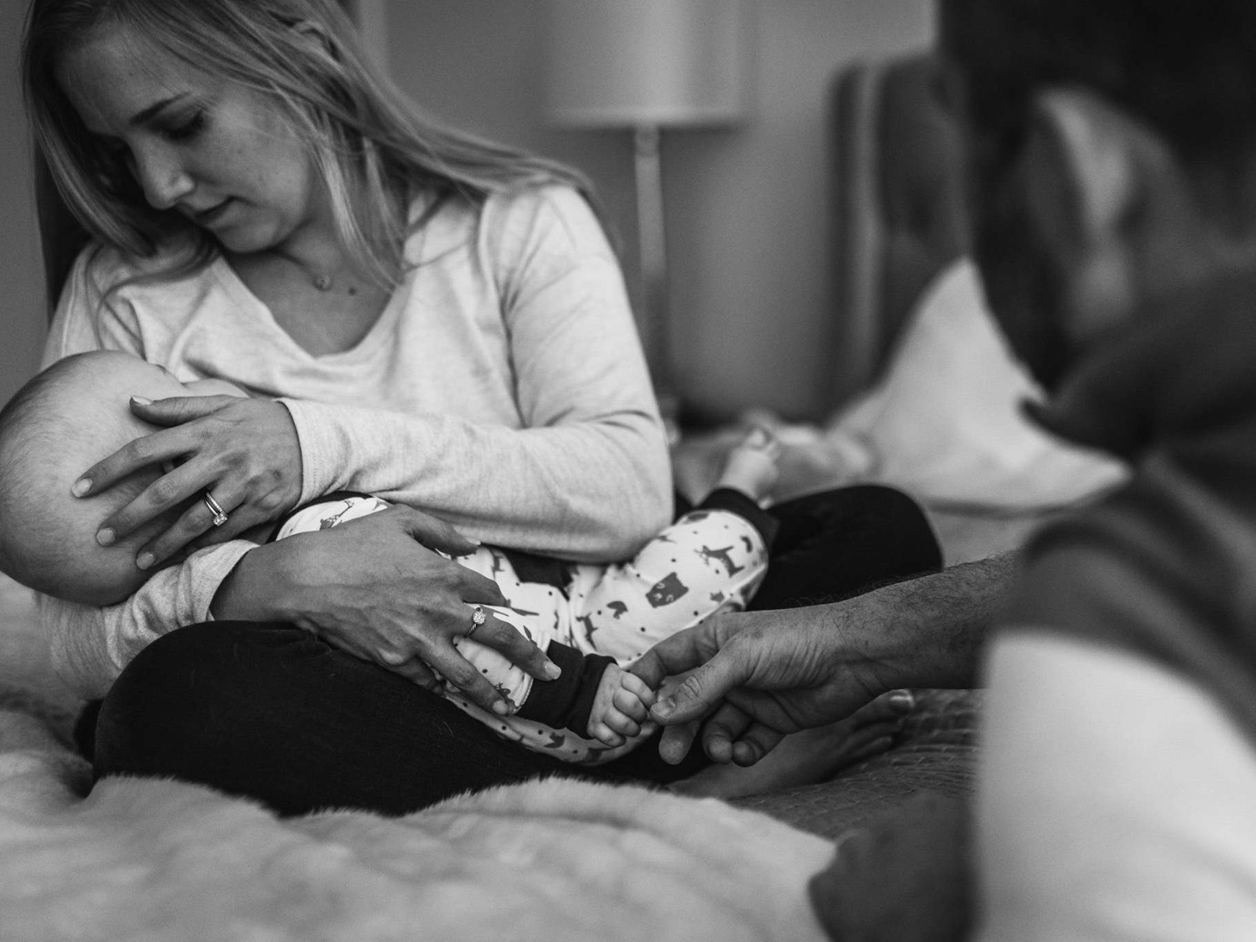 Ellen gazing down at newborn Gavin in her arms, black and white documentary photo. A second pair of hands reaches in to hold his feet.